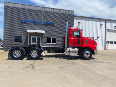 Used Mack truck with red cab and long chassis in front of Used Truck Center building. Visible text includes 'USED TRUCK CENTER' on the building, and no other legible labels on truck.