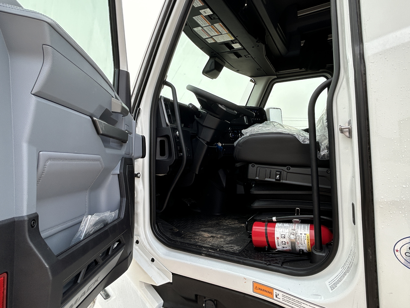 Truck cab interior view of a 2026 Mack PR64T44 showing driver seat, dashboard, and open door with fire extinguisher mounted on floor.