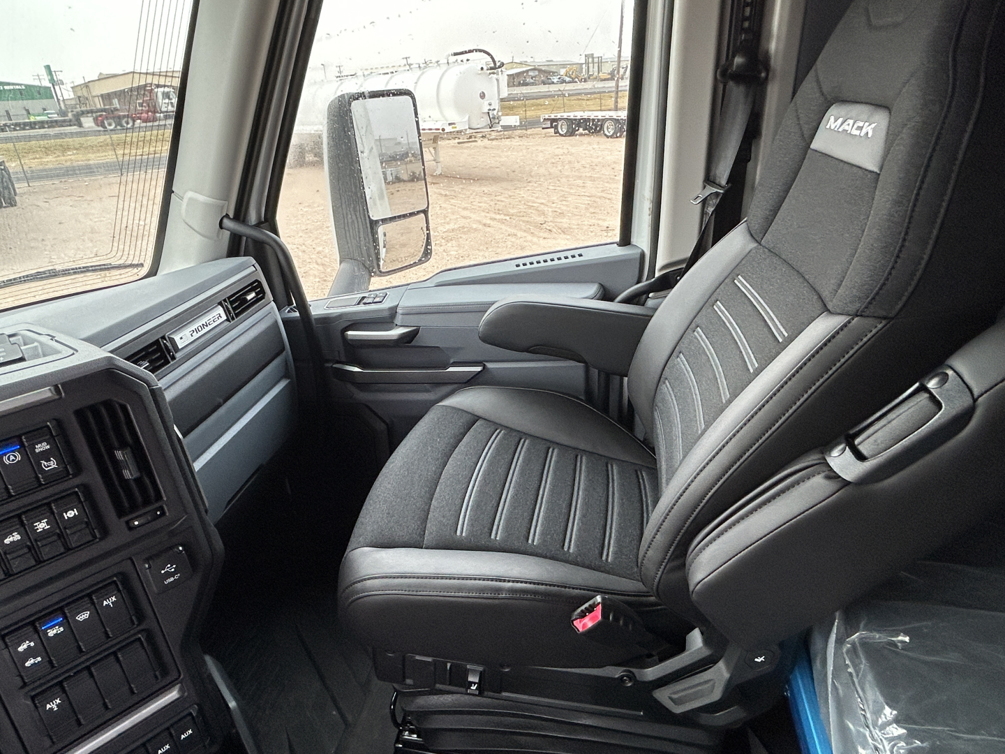 Truck cabin interior showing driver seat with Mack logo, dashboard controls, and side window view.