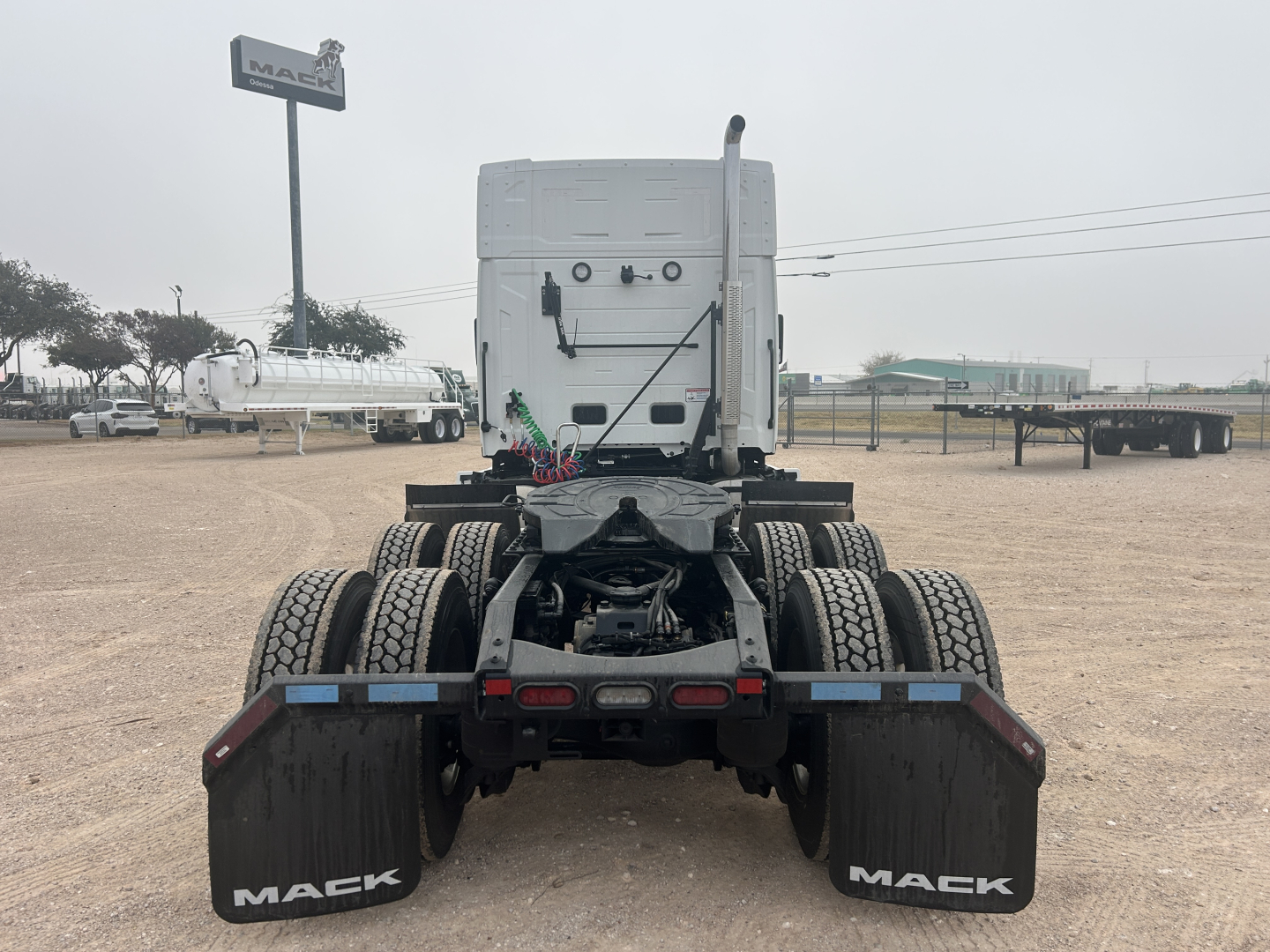 Rear view of a new Mack PR64T44 truck chassis showing dual rear axles and mud flaps with Mack logo, in a dealership lot.