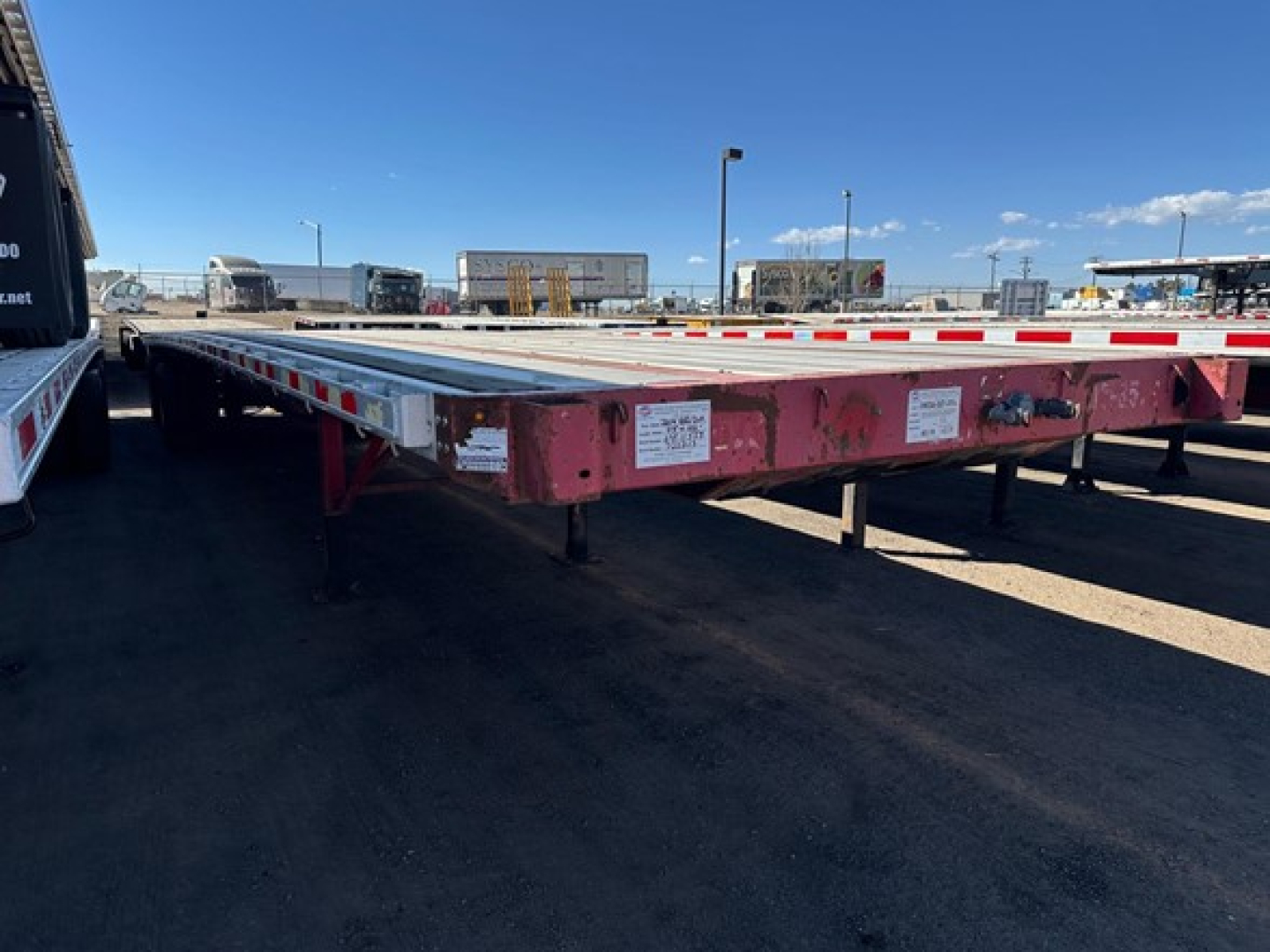 Flatbed trailer showing long red frame and white deck with multiple corner and side labels, parked at a yard.