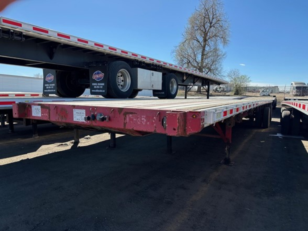 Flatbed trailer with a raised gooseneck and a long deck, Great Dane branding on front ramps, undercarriage showing tires and support legs.