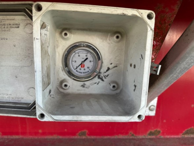 Gauge in a metal recessed box on red trailer body. Circular dial with numbers from 0 to 60, red needle, mounted in square, weathered housing with screws at corners.