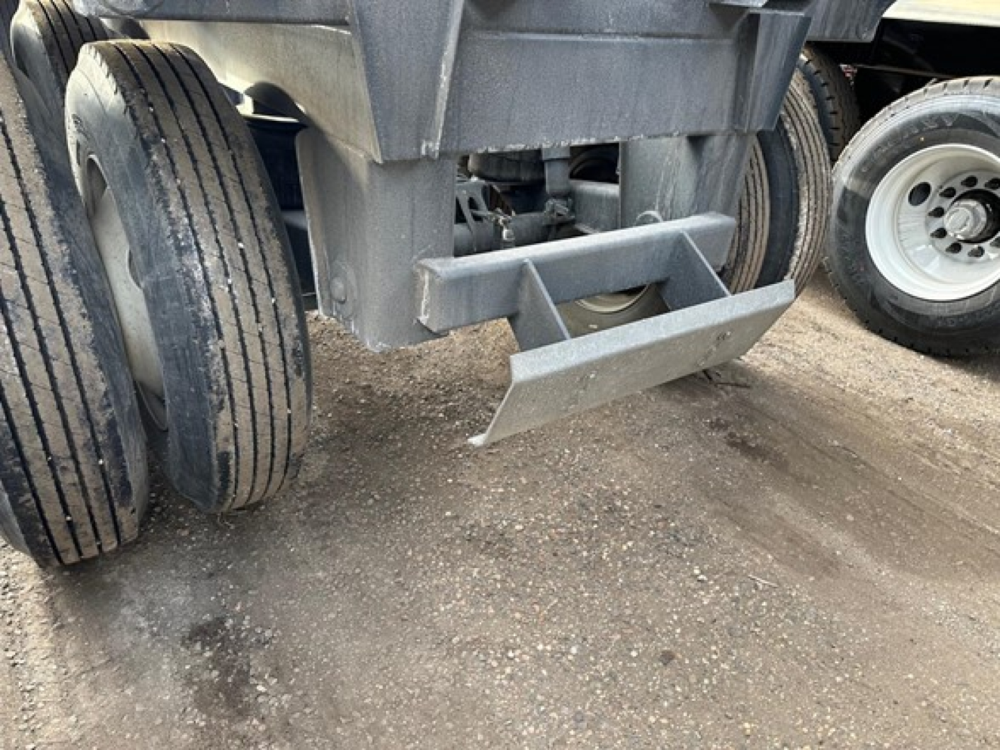 Truck trailer hitch and rear underside showing dual rear axles and dump body, with mud/grime on ground.