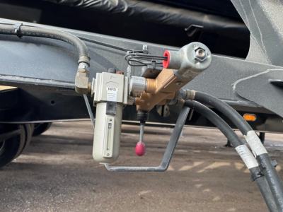 Trailer hydraulic manifold with pump and hoses showing fittings and a small vertical reservoir cap, attached to a frame member.