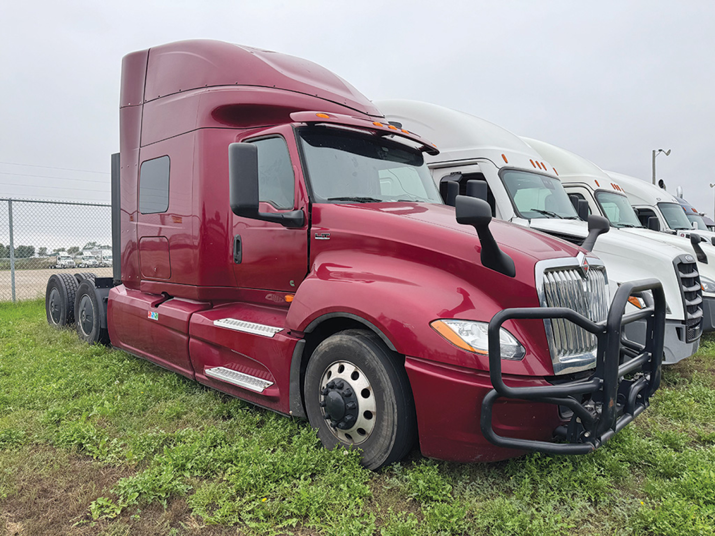 Truck cab exterior of a red 2021 International LT625 sleeper tractor, side view with front grille guard and black mirrors, other trucks in background.