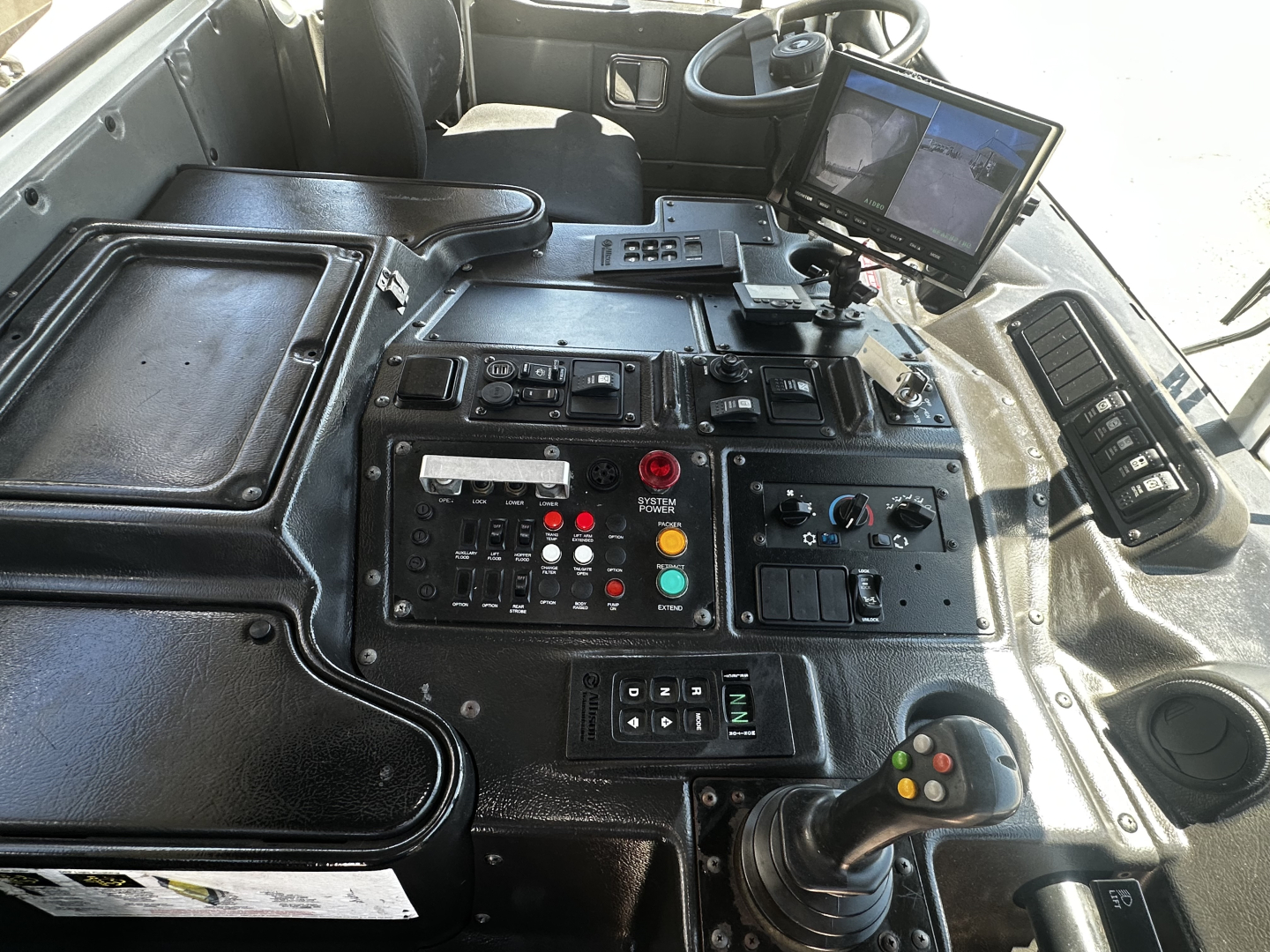 Dashboard view of a vehicle cab with control panel, joystick, touchscreen monitor, and multiple switches labeled with small text visible.