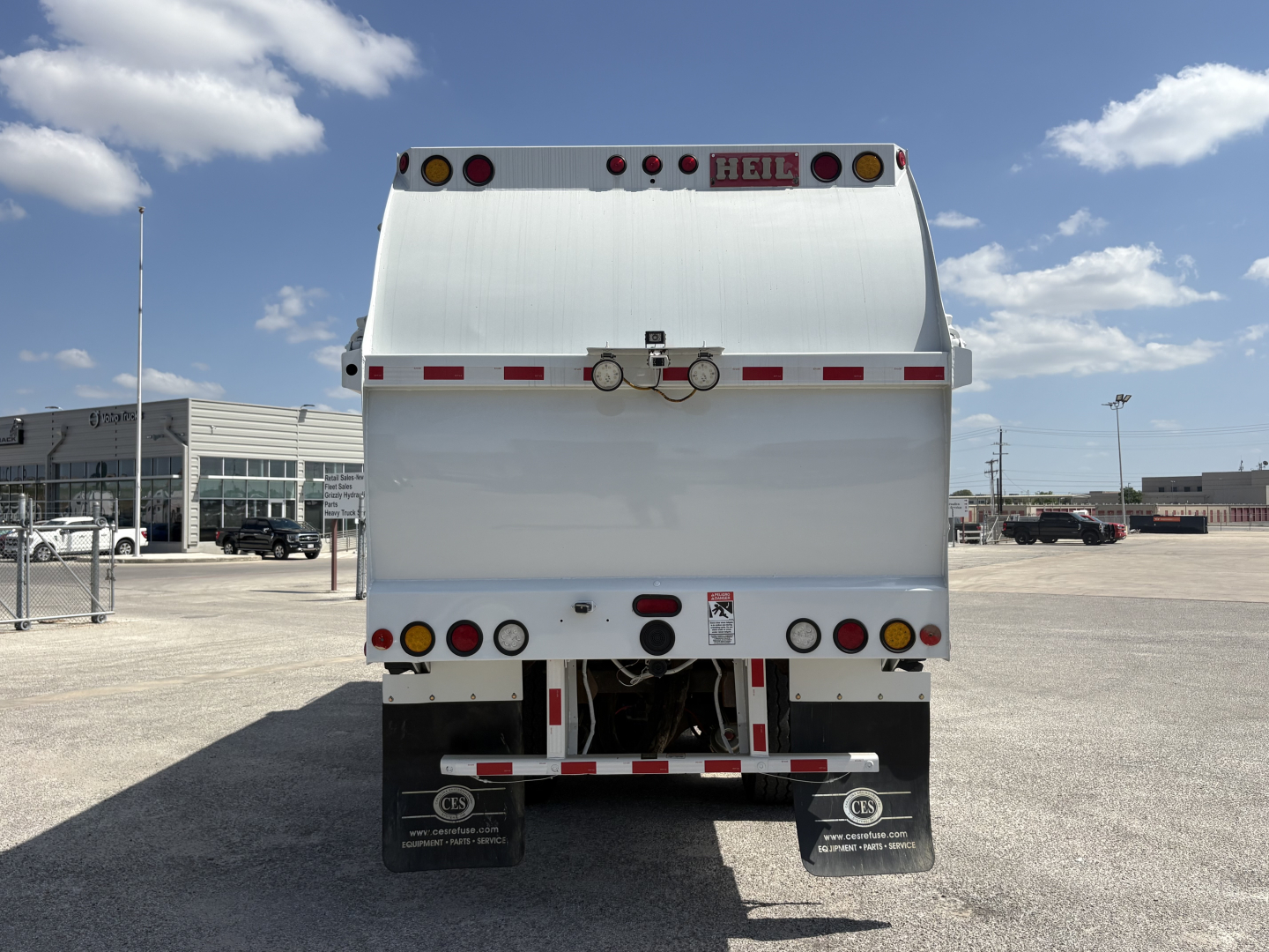 Rear view of a white Heil garbage truck body with taillights, reflective strips, and a hydraulic apparatus; text includes 'HEIL' on top center and bumper area details.