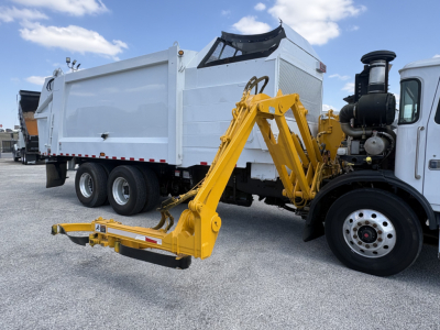 Autocar XPEDITOR truck with a yellow hydraulic arm attachment connected to the front. White dump body in background, black exhaust stack, and twin rear axles visible.