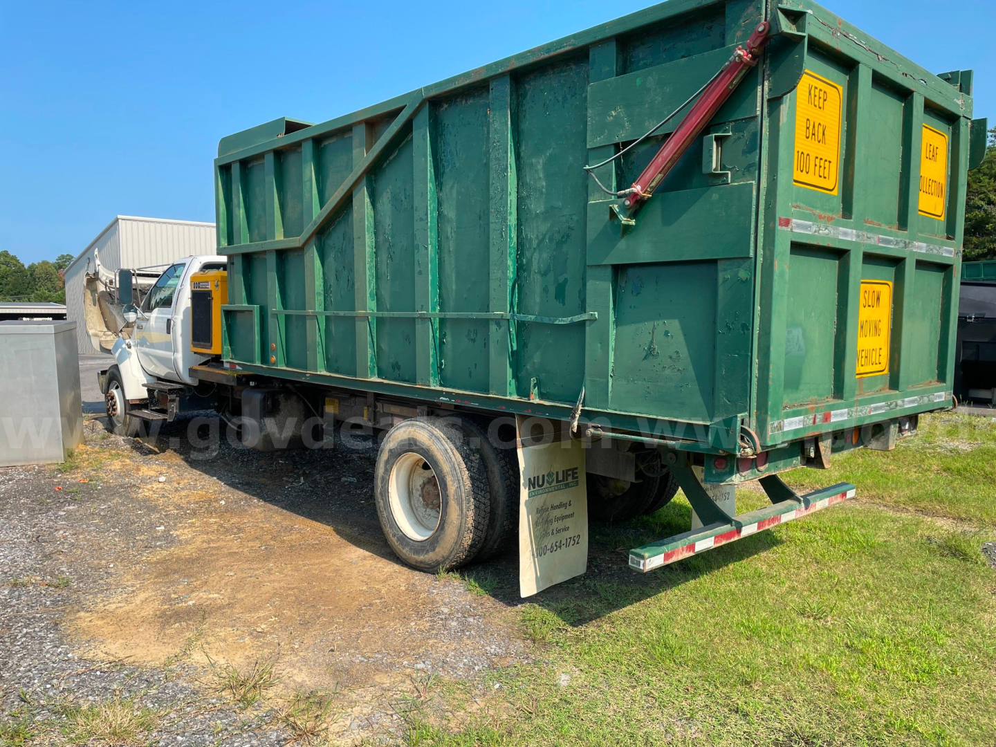 Truck bed with a large green waste container on a Ford F-750 chassis, side view showing yellow warning labels and rear bumper.