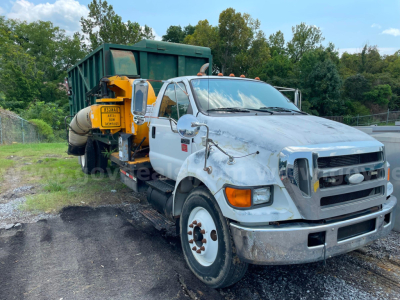 Ford F-750 vacuum service truck with white cab and front bumper, yellow mounted vacuum unit behind the cab, and green dump body in backdrop.