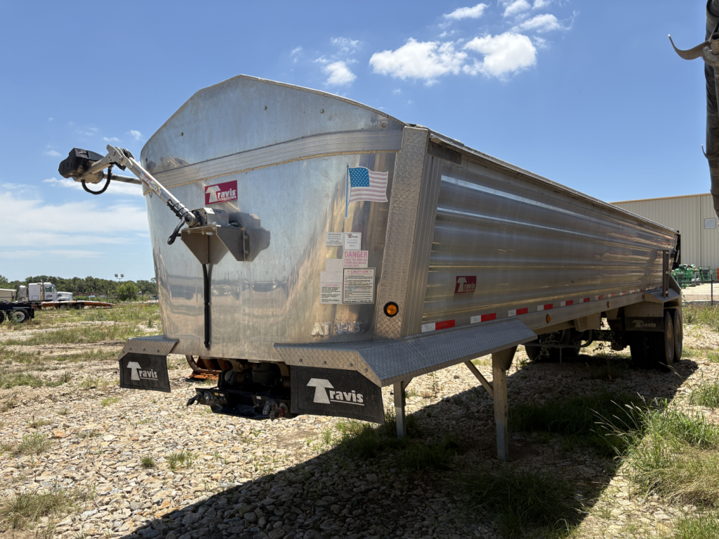Trailer side and front of a 2024 Travis Wave dump trailer with a front hitch, metal side panels, and a small American flag sticker on the side.