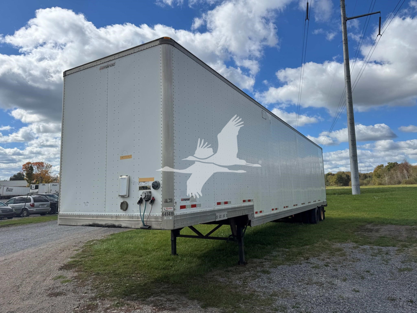 Trailer side view showing a 2015 Manac drop van with a white logo on the side, rear access, and landing legs visible in a grassy area.