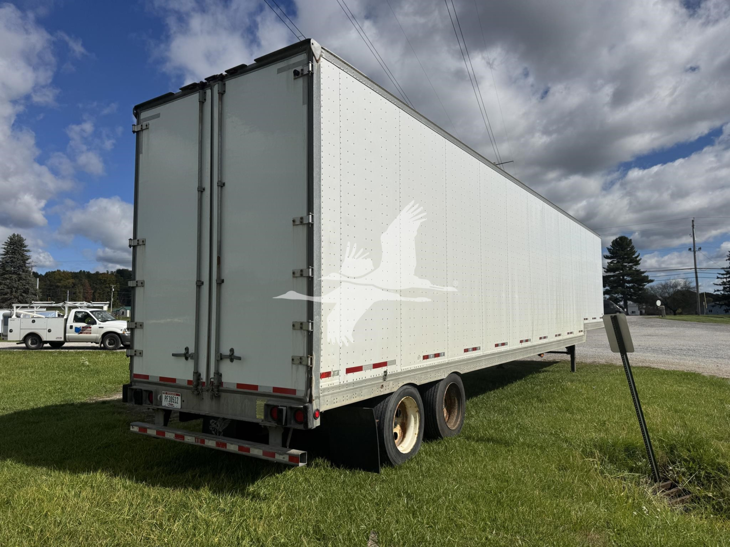 Manac drop van trailer viewed from rear-left angle showing white cargo box, dual rear doors, and tandem axles on grassy lot.