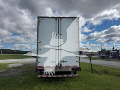 Rear view of a Manac sheet and post trailer with dual rear doors and reflective tape, standing on grass with other trailers in distance.