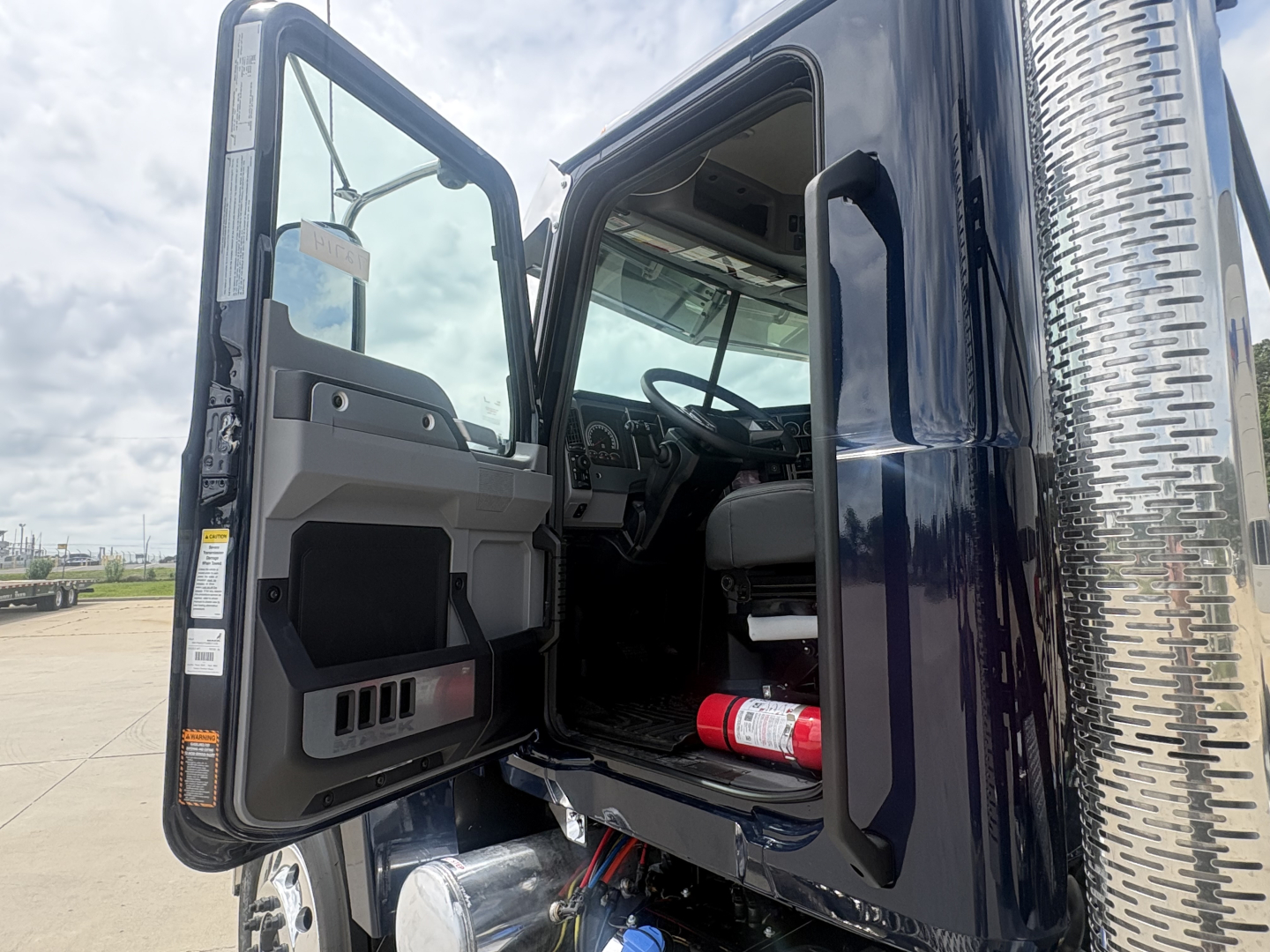 Truck cab interior and open door showing steering wheel, dashboard, and side step; is a new 2027 Mack PI64T conventional daycab with chrome exhaust stack visible on right.