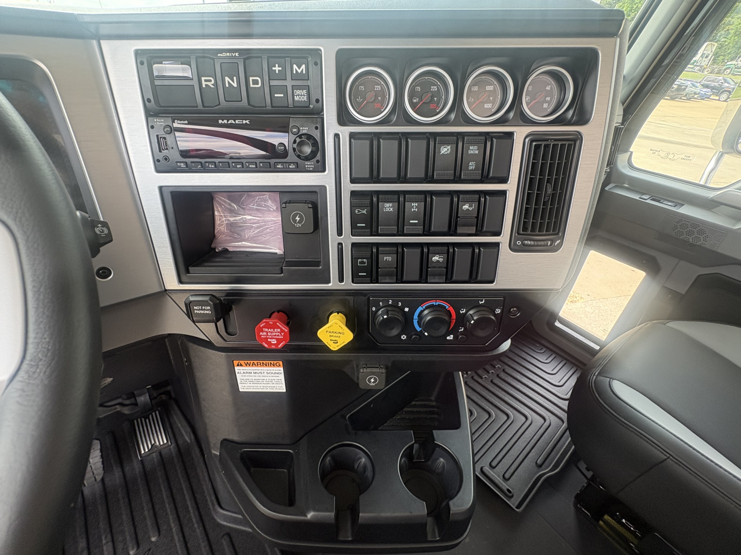 Dashboard view of a Mack PI64T truck cab showing gear selector, gauge cluster, climate controls, cup holders, and switch panel.
