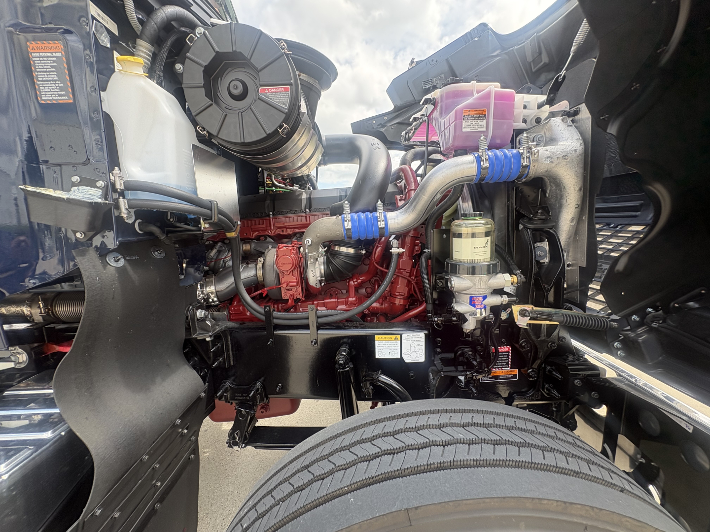 Mack PI64T engine bay showing intake, coolant reservoir, pink coolant overflow, hoses, and blue hose clamps near front wheel.