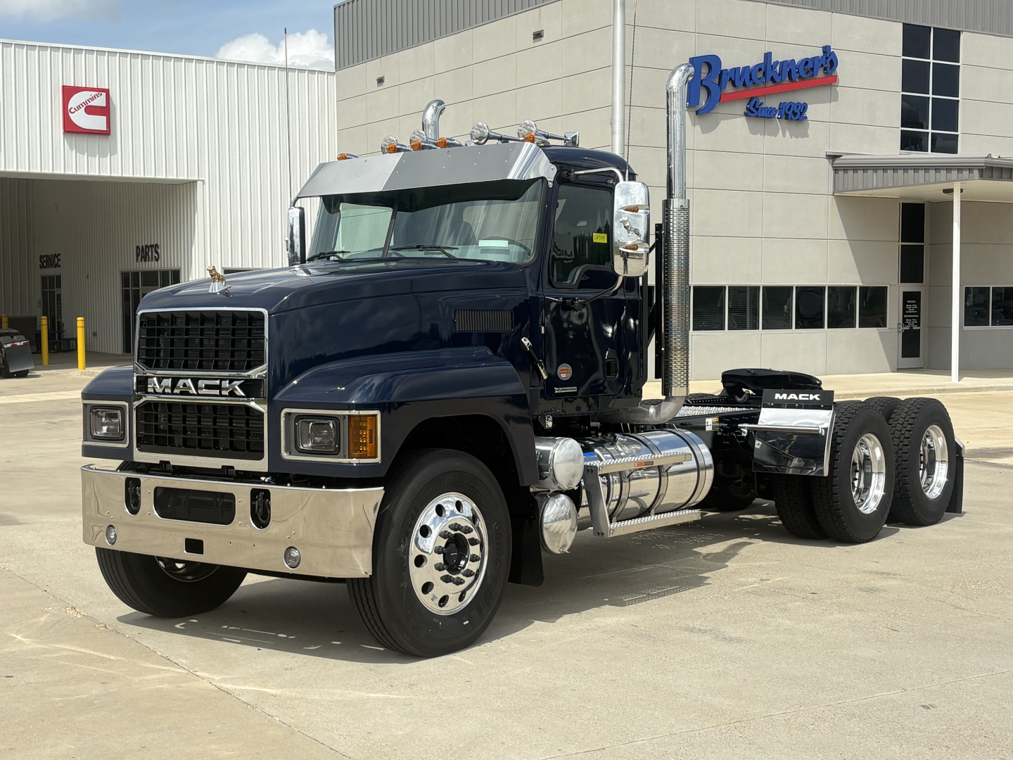 Mack PI64T conventional day cab truck showing front grille, chrome bumper, dual exhaust stacks, fuel tanks, and triple axle configuration.