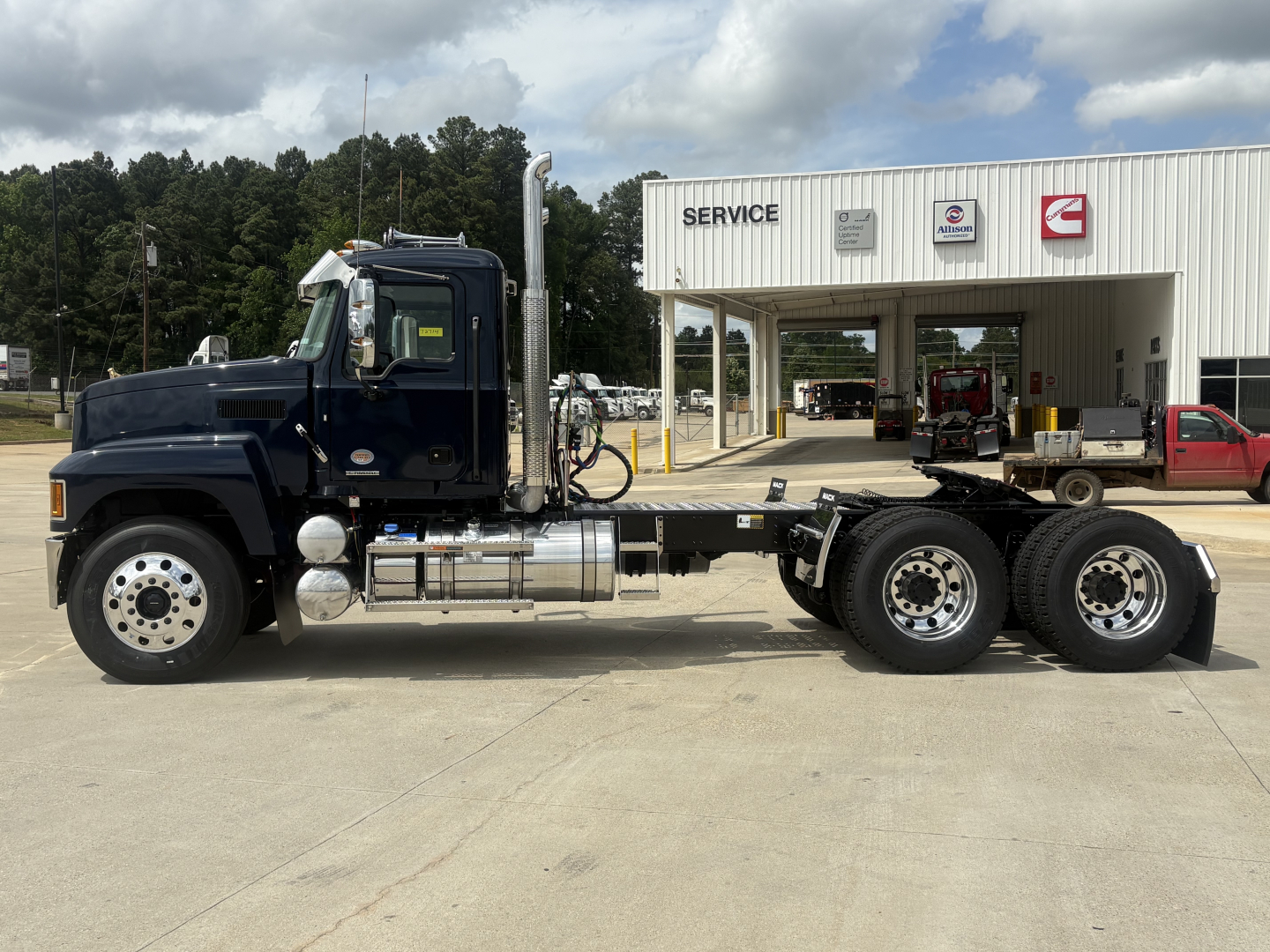 Truck: Mack PI64T conventional daycab chassis with cab and dual rear axles in front of service building; visible tanks and exhaust stacks; no background text readable other than signs on building.