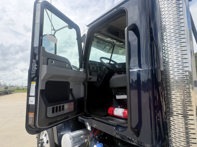Truck cab interior and open door showing steering wheel, dashboard, and side step; is a new 2027 Mack PI64T conventional daycab with chrome exhaust stack visible on right.