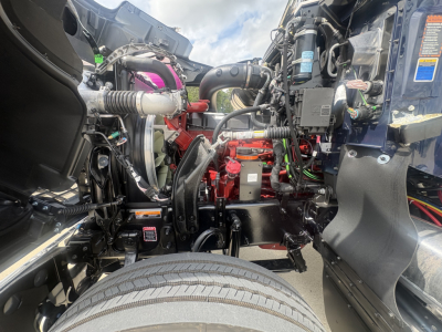 Mack PI64T engine bay showing red engine block with surrounding hoses and wiring, front wheel in foreground, various components and label stickers visible.