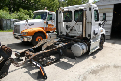 Truck chassis and cab with Freightliner Cascadia 113 truck in background. Front of white cab shows exposed drivetrain; orange-and-white truck in background with numbers on its door.