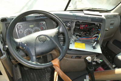 Cab interior of a 2014 Freightliner Cascadia 113 showing steering wheel, dashboard, exposed wiring, and dash area with a yellow tag on a metal panel.