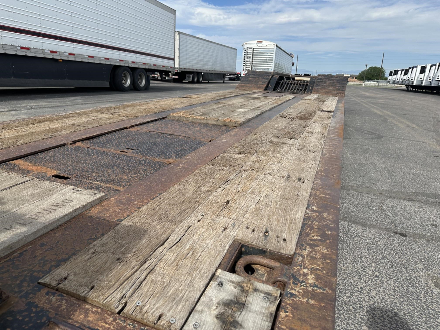Trailer bed with weathered wooden deck and rusted steel edges, long lowboy trailer in an industrial lot, showing loading ramps and tie-down anchors.