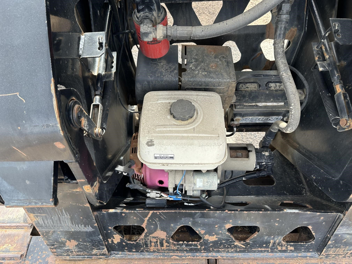 Small engine compartment of a Trail King lowboy trailer showing a gray starter/generator housing, pink fuel line, black battery box, and surrounding metal frame with dirt.