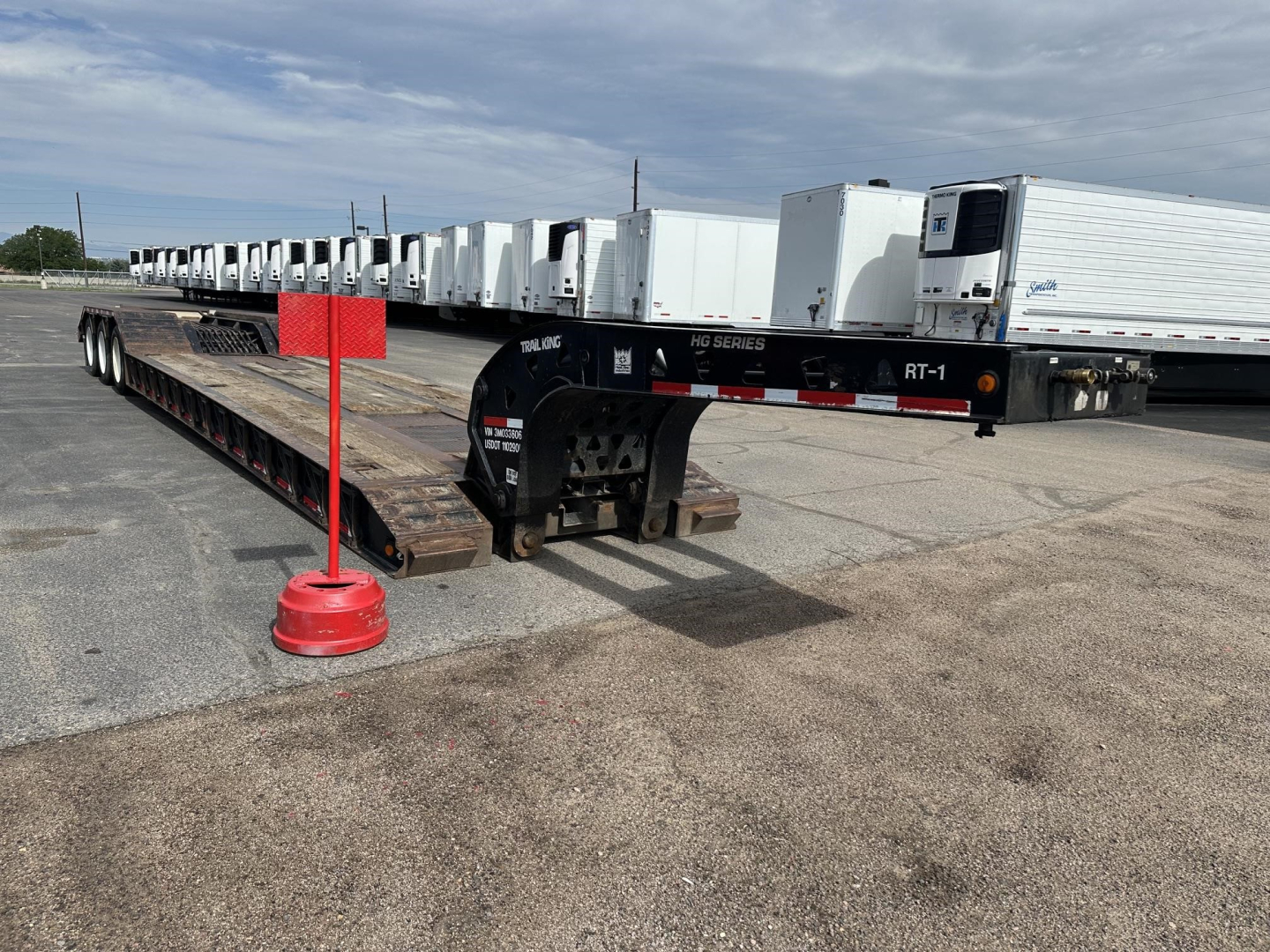 Trail King lowboy trailer with long flatbed and gooseneck. Row of white trailers in background; red pole marker and black gooseneck neck with RT-1 and HG Series markings visible.