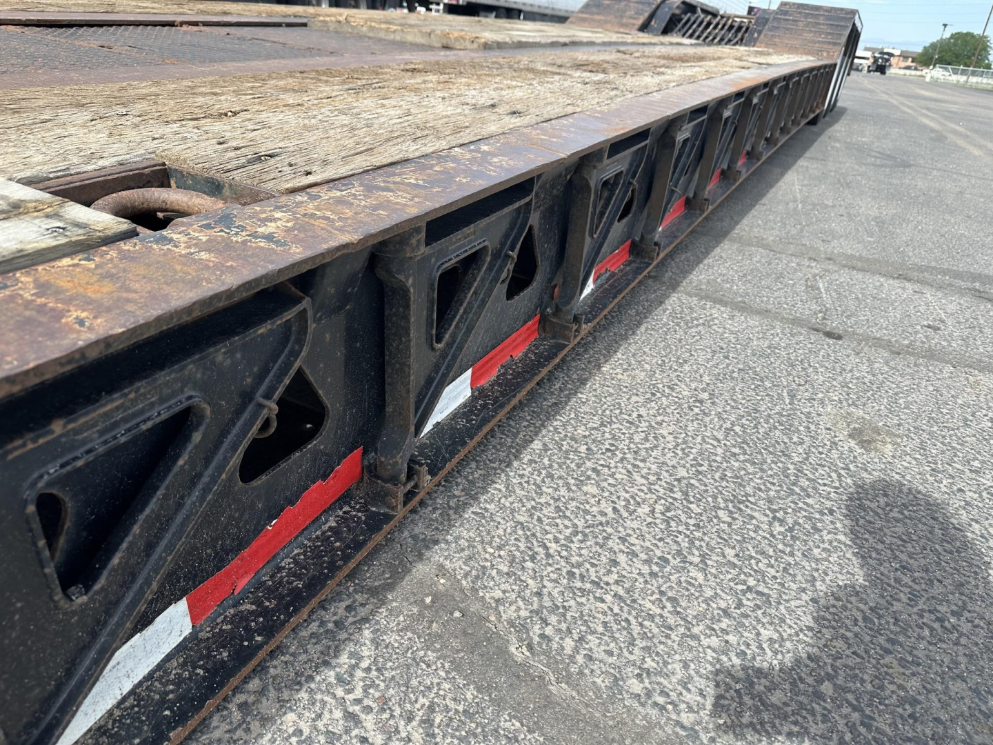 Lowboy trailer side with black structural frames and red/white reflective tape along the edge. Long flatdeck with wooden surface and rust on steel components.