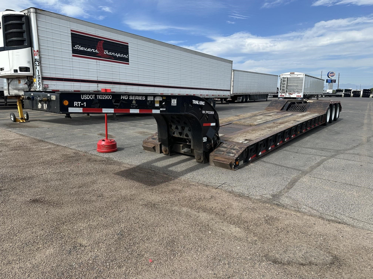Lowboy trailer with deck and tandem axles parked in lot; visible branding on gooseneck and white refrigerated trailer in background; USDOT 902180 label and Series markings.