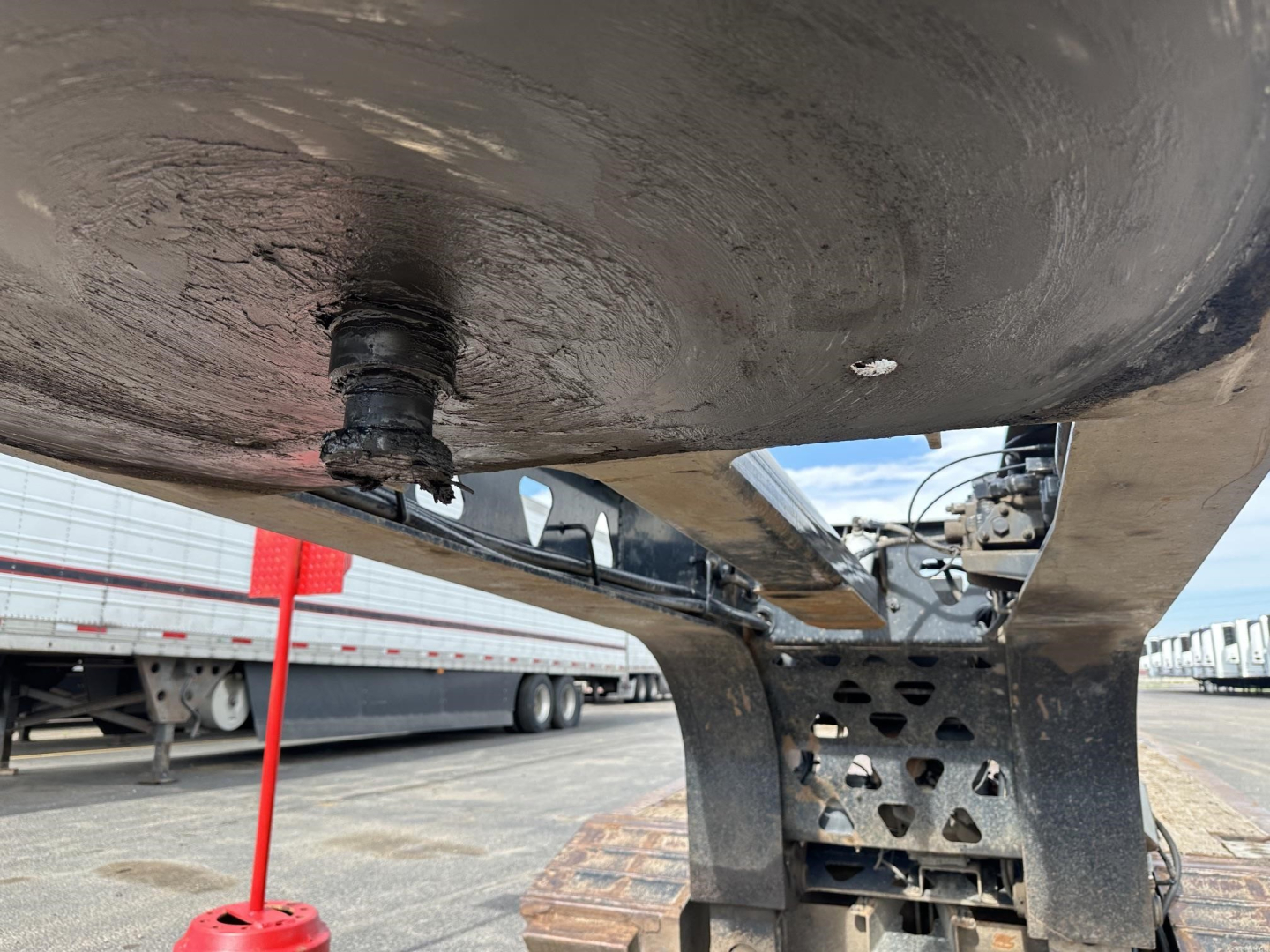 Lowboy trailer detail showing mounting hardware and underside of a round tank or deck with bolted fitting and reinforced steel frame, surface grime present.