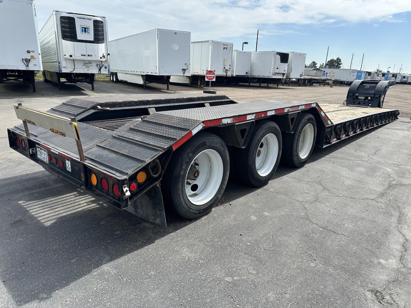 Trailer showing a long lowboy with triple axles and a rear access ramp, black metal deck, and white wheels in an outdoor lot.