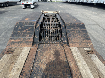 Heavy rusted lowboy trailer with open burn pit and grated center, viewed from the front showing the deck and wheel wells.