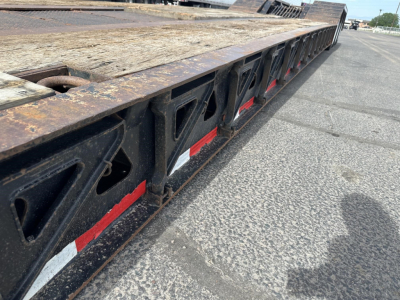 Lowboy trailer side with black structural frames and red/white reflective tape along the edge. Long flatdeck with wooden surface and rust on steel components.