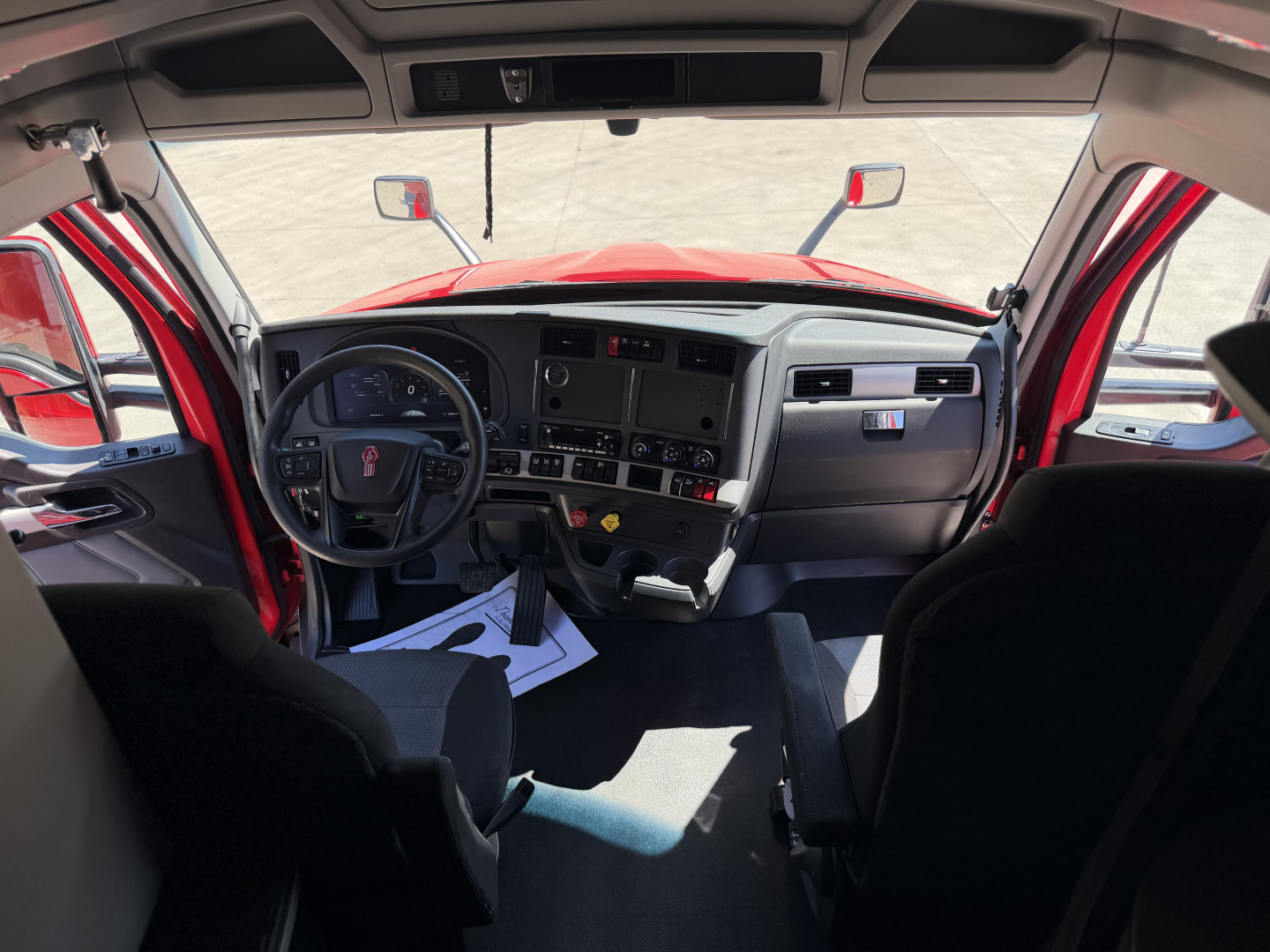 Truck cab interior of a 2024 Kenworth T680 showing dashboard, steering wheel, and driver seat with paperwork on floor