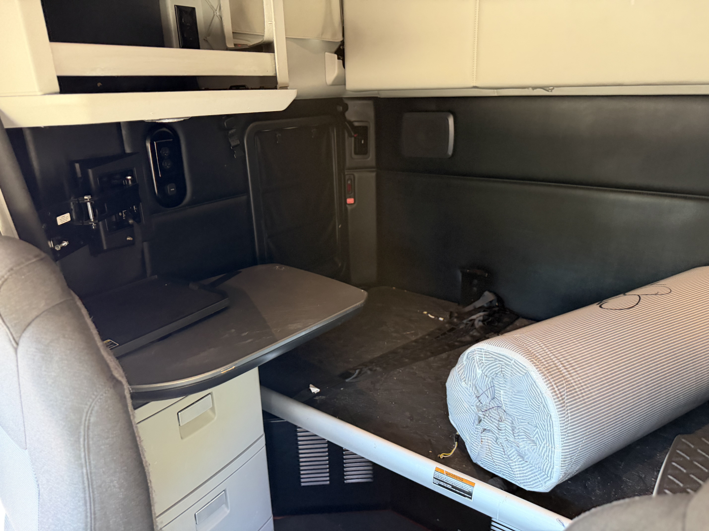 Kenworth T680 cab interior showing sleeper area with a fold-out table, storage cabinet, and rolled foam mattress near a passenger seat.