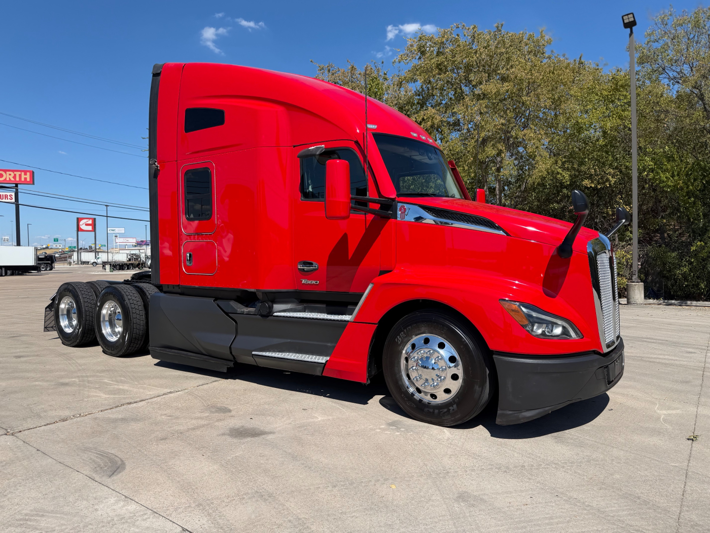 Kenworth T680 tractor in red, side view showing cab, chrome wheels, two rear axles, and aerodynamic mirrors.