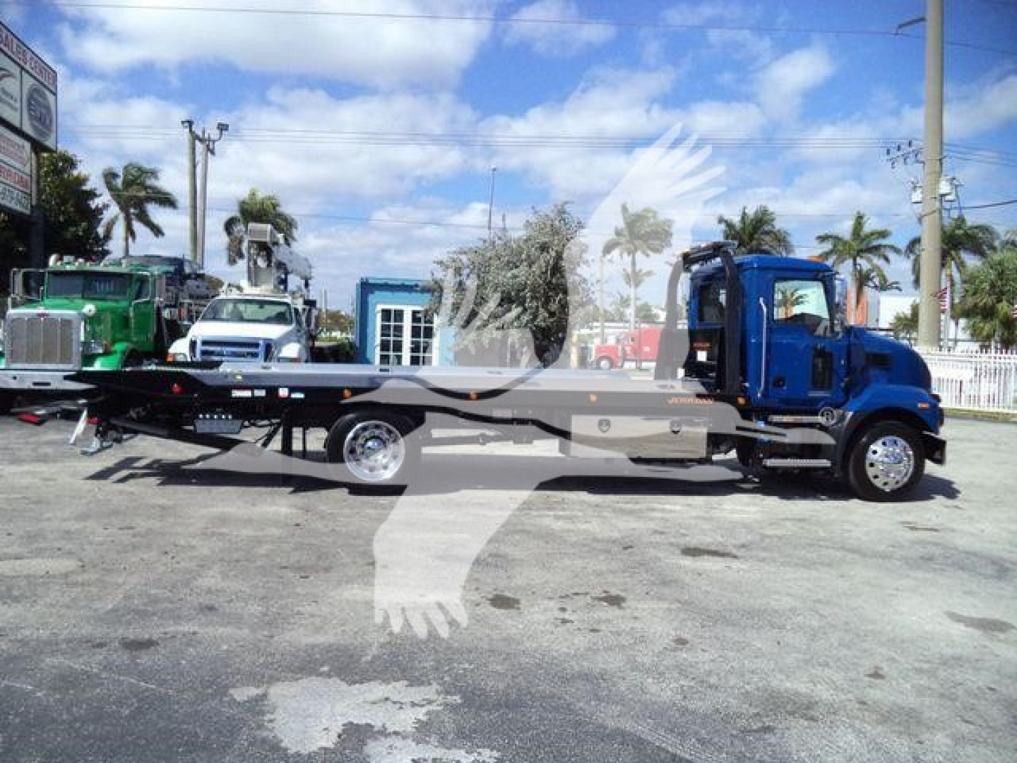 2027 Mack MD6 rollback truck side view showing flatbed and blue cab with other trucks in background