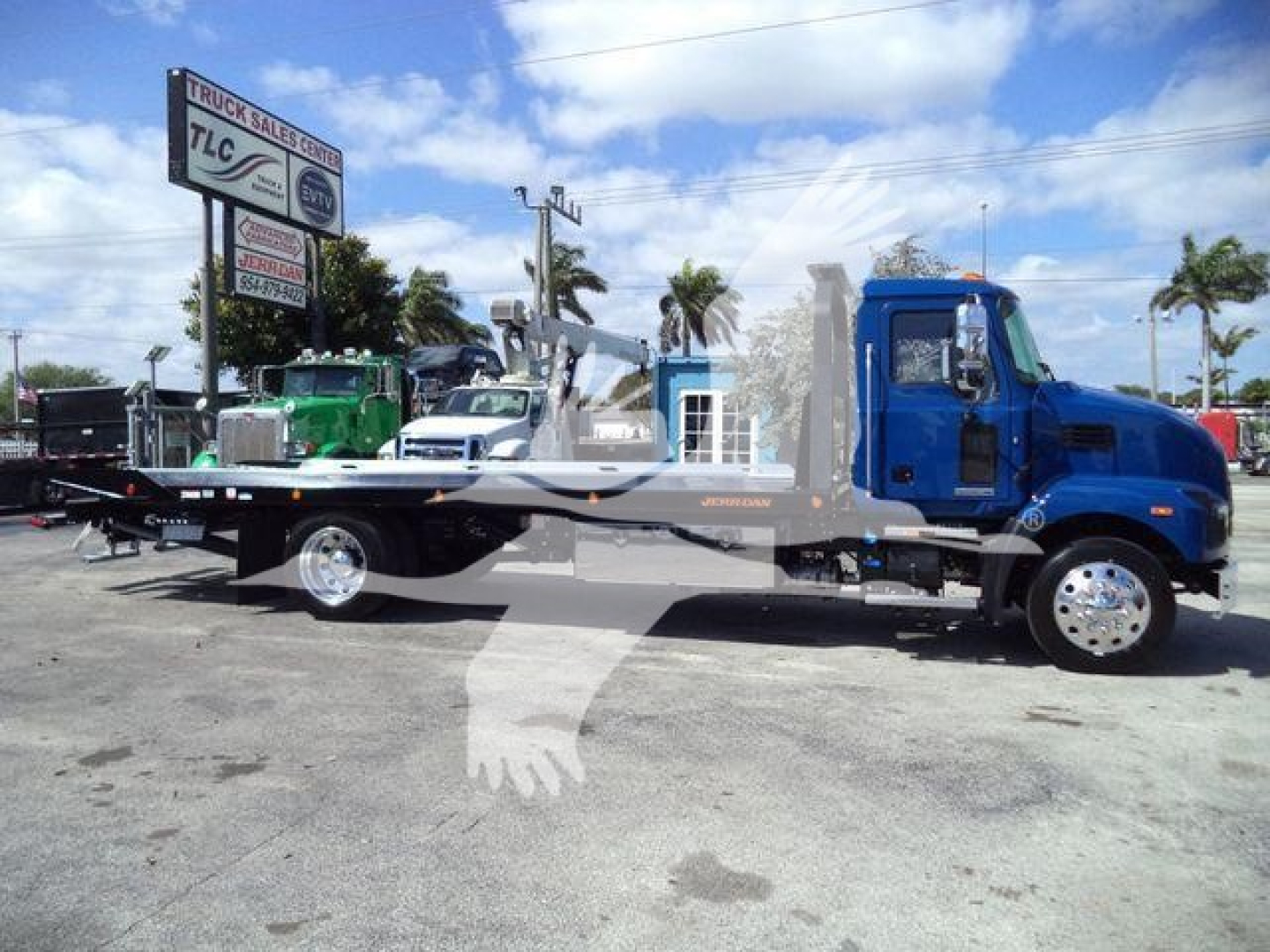 Blue Mack MD6 rollback tow truck with long flatbed and chrome wheels, parked at a dealership lot.