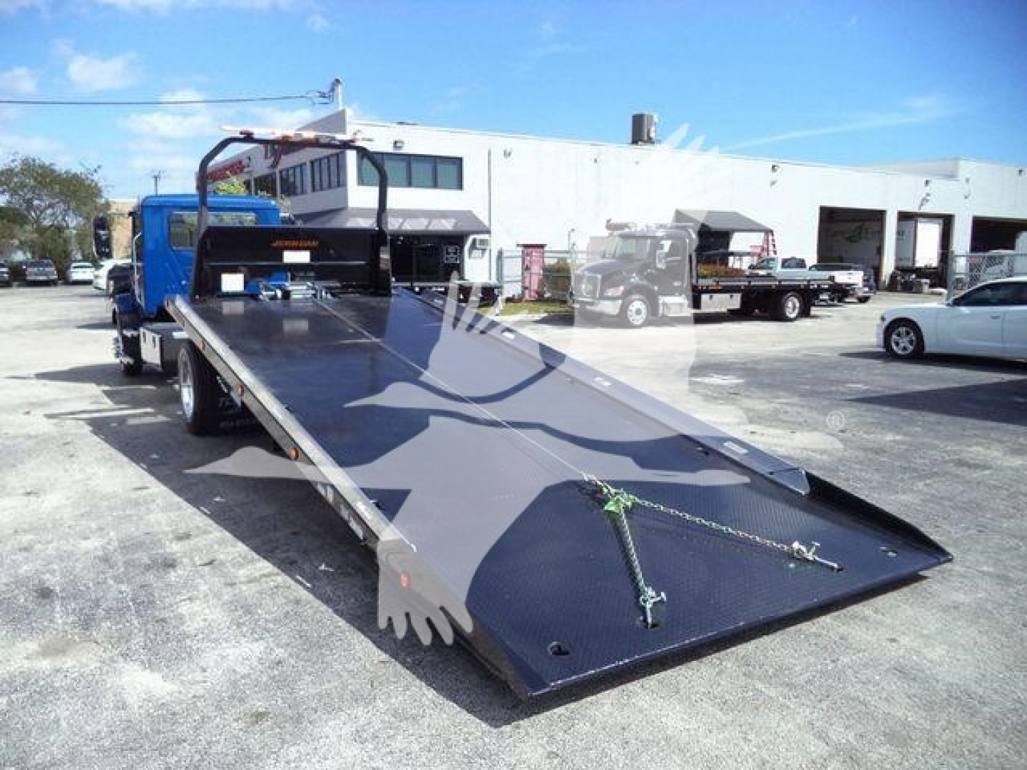 MD6 rollback tow truck with a flatbed in lowered position, blue cab, white trailer in background, open parking lot.