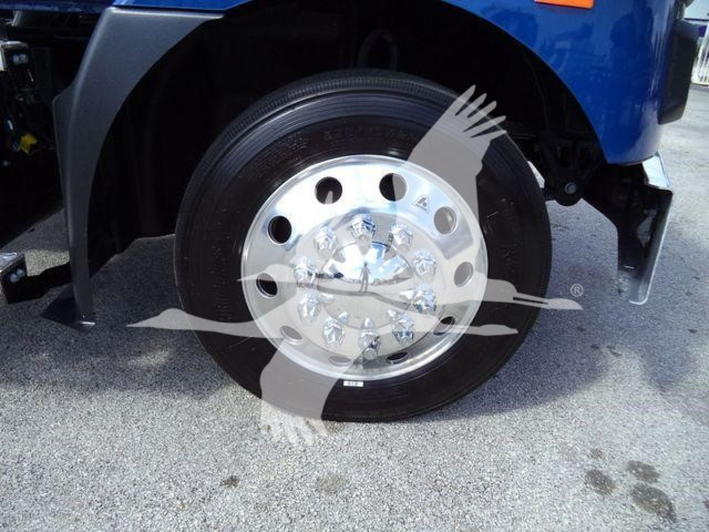 Front wheel of a blue Mack MD6 truck showing polished wheel with lug nuts and tire.