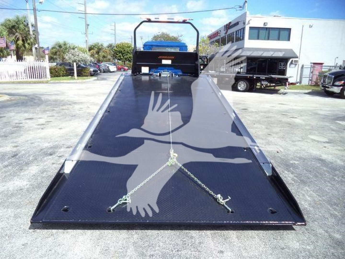 Mack MD6 rollback truck with a wide lowered flatbed ramp in front, black with chain tie-downs, rear photo shows loading area and wheel fender in background.