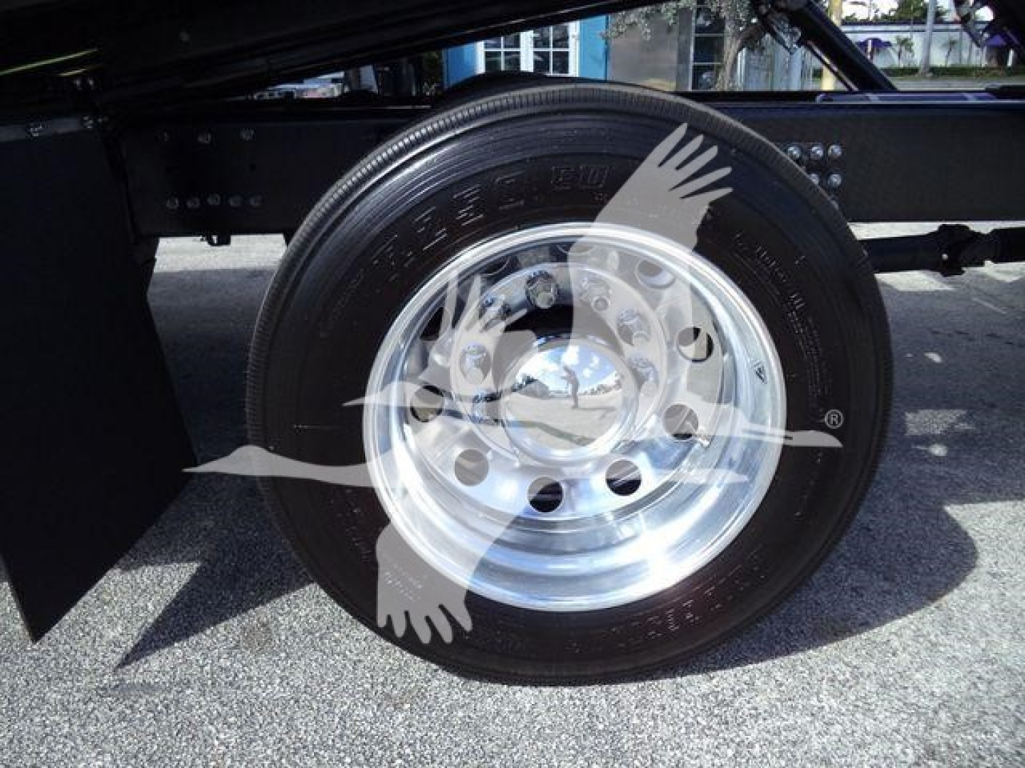 Front wheel and tire assembly on a Mack MD6 rollback truck showing polished steel wheel and black tire with visible tread, undercarriage components in background.