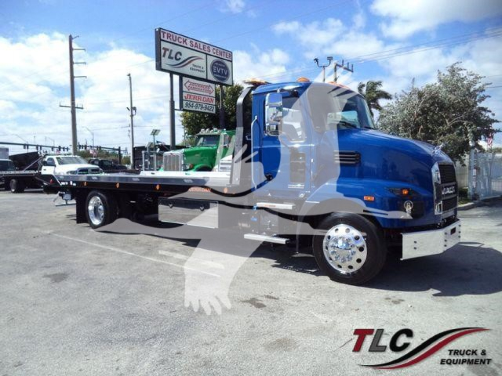 Blue Mack MD6 rollback truck in showroom lot with long flatbed, chrome wheels, and TLC Truck Sales Center sign in background.