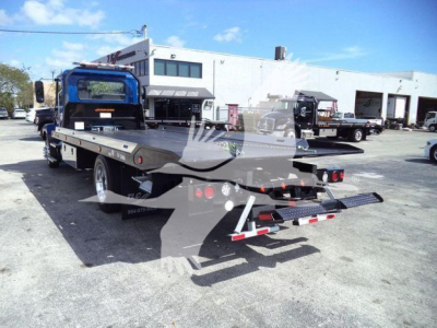 Flatbed tow truck in a lot with a blue cab and a white building in background. Flatbed shows warning lights and rear loading ramps.