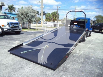 Mack MD6 rollback truck with a long black flatbed in a parking lot, showing front ramp surface and metal tie-downs.