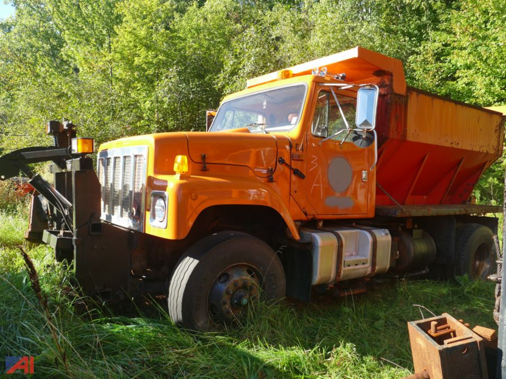 Orange 1997 International 2574 dump truck with sander attachment, parked among grass and trees.